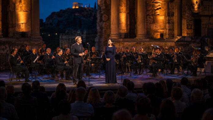 agia-paraskevi-bezos-tsaliki-filharmoniki-20260824-1630-1920x1080-1 Δύο ερμηνευτές μπροστά σε ορχήστρα πνευστών σε βραδινό υπαίθριο θέατρο