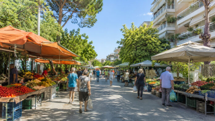 Υπαίθρια λαϊκή αγορά με πάγκους οπωροκηπευτικών σε δενδροφυτεμένο αστικό δρόμο