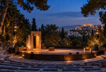 Γαλάτσι: Απόψε η παράσταση «Όπου γης και Patris» στο Άλσος Βεΐκου Φωτισμένη υπαίθρια θεατρική σκηνή με βαλίτσα και καρέκλα σε καταπράσινο αθηναϊκό τοπίο