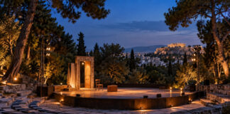 Γαλάτσι: Απόψε η παράσταση «Όπου γης και Patris» στο Άλσος Βεΐκου Φωτισμένη υπαίθρια θεατρική σκηνή με βαλίτσα και καρέκλα σε καταπράσινο αθηναϊκό τοπίο