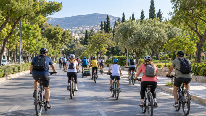 Οικογένειες και ενήλικες ποδηλάτες με κράνη κινούνται σε δενδροφυτεμένο δρόμο