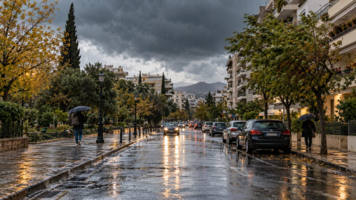 Βροχερός δρόμος σε αστική γειτονιά της Αττικής με πεζούς που κρατούν ομπρέλες